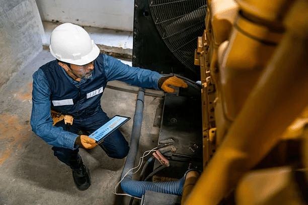 Technician in a hard hat inspecting a commercial standby generator and recording readings on a tablet
